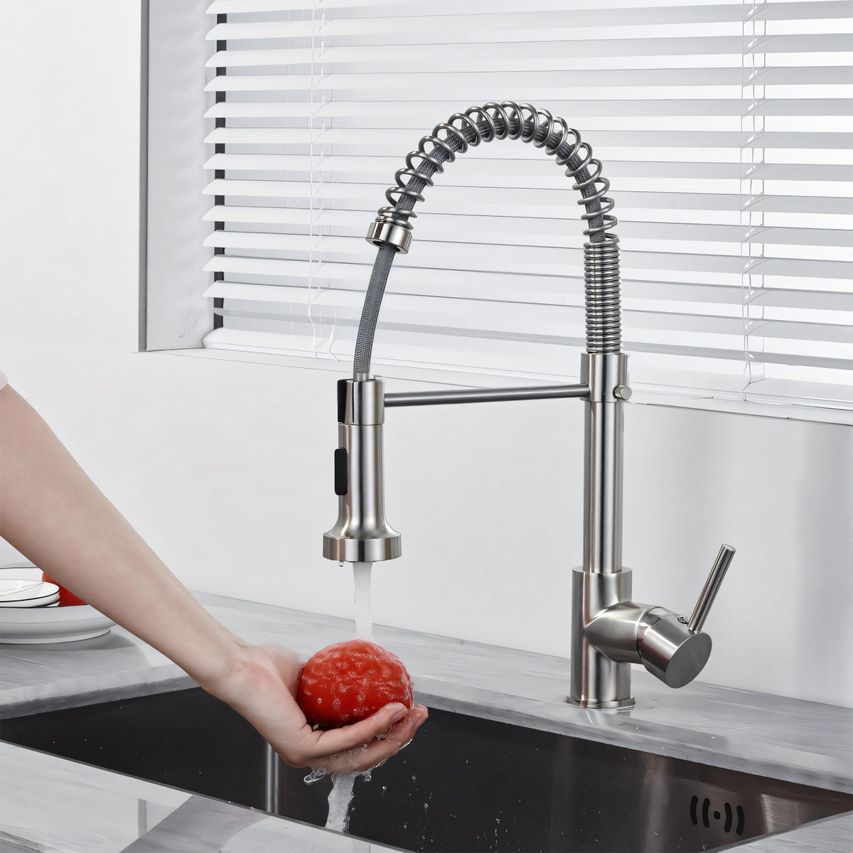 A modern pull-down kitchen faucet with a flexible spring coil neck and single lever handle, shown in a sink with water running while a hand holds a red tomato. The faucet has a brushed silver/chrome finish.